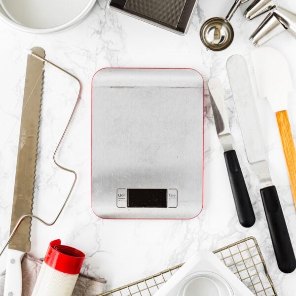 A curated collection of essential tools for egg-free baking is artfully arranged on a marble surface, all centered around a digital kitchen scale.