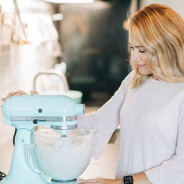 A woman operates a teal stand mixer in a kitchen, exploring her top tips for baking without eggs. She wears a light pink blouse and a smartwatch, with her light brown hair styled loose. The kitchen features wooden cabinets and a tiled backsplash.