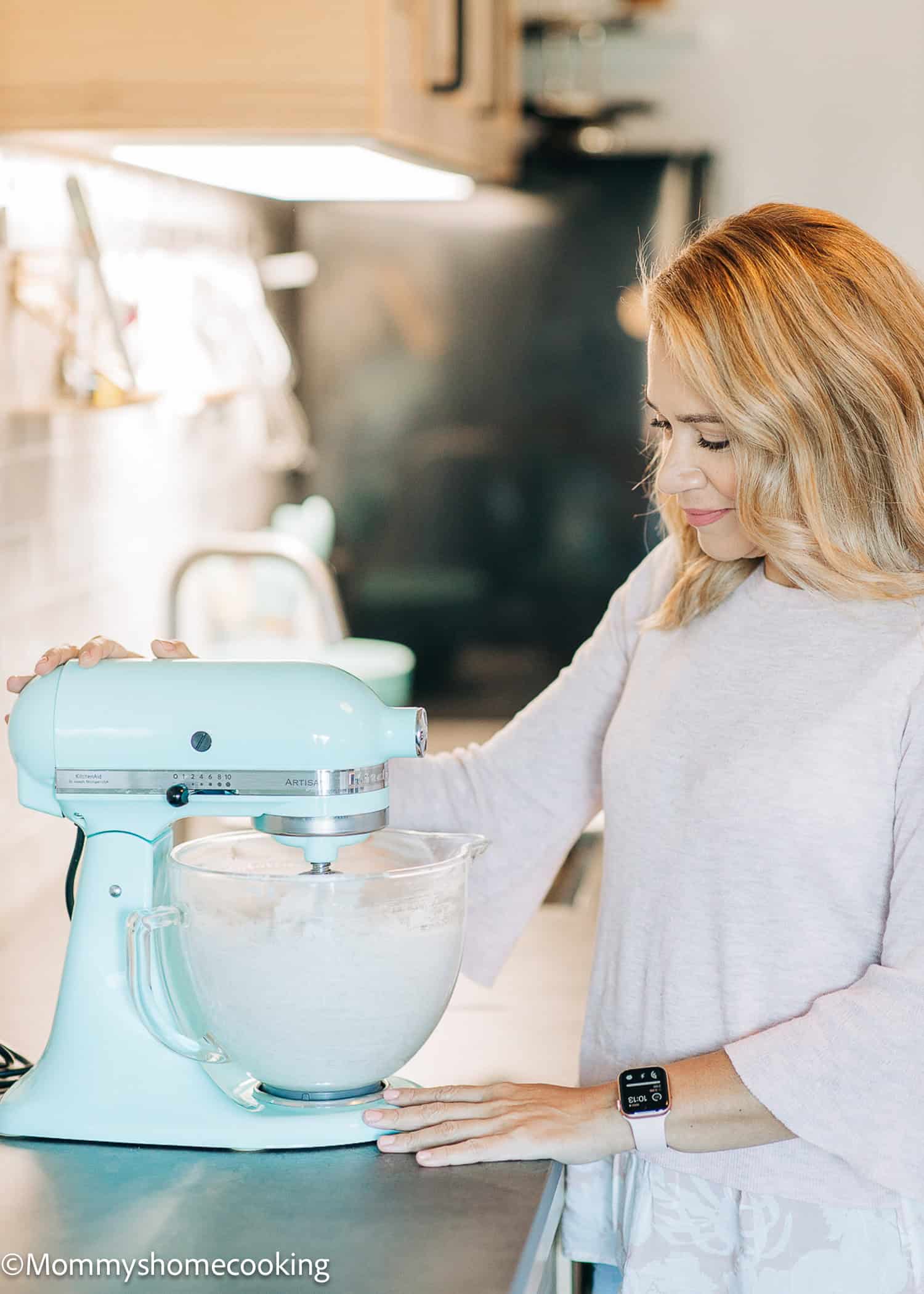 A woman operates a teal stand mixer in a kitchen, exploring her top tips for baking without eggs. She wears a light pink blouse and a smartwatch, with her light brown hair styled loose. The kitchen features wooden cabinets and a tiled backsplash.