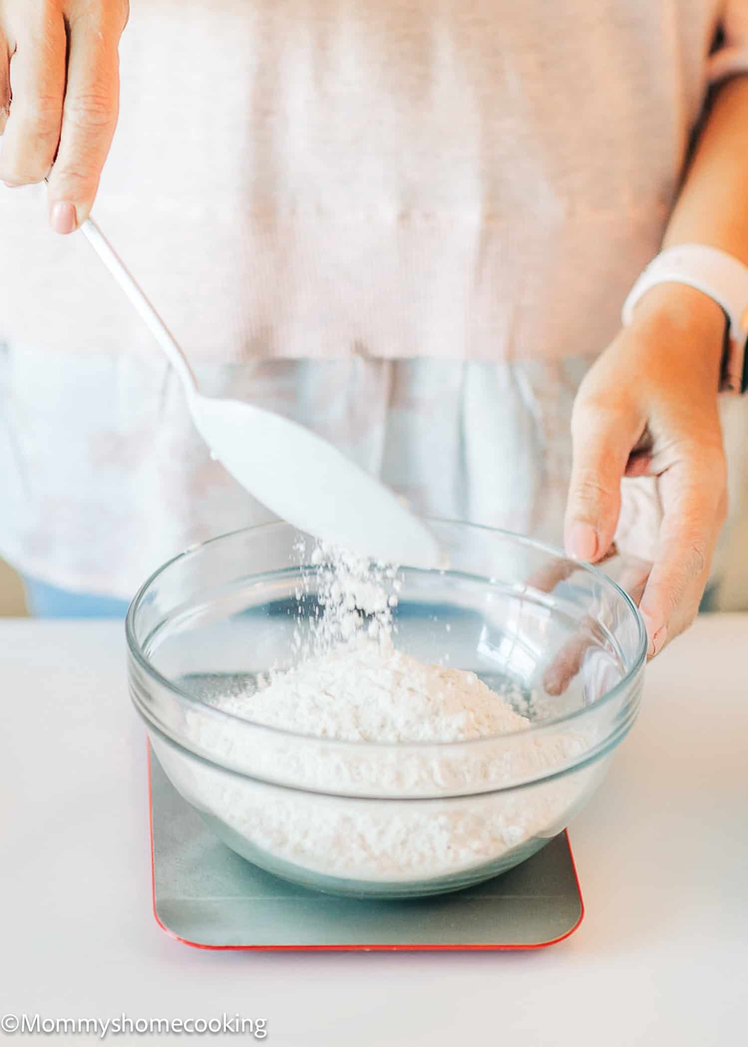 A person carefully adds flour from a spoon into a glass bowl already filled with flour, following essential steps from The Complete Guide to Measuring and Mixing for Egg-Free Baking.
