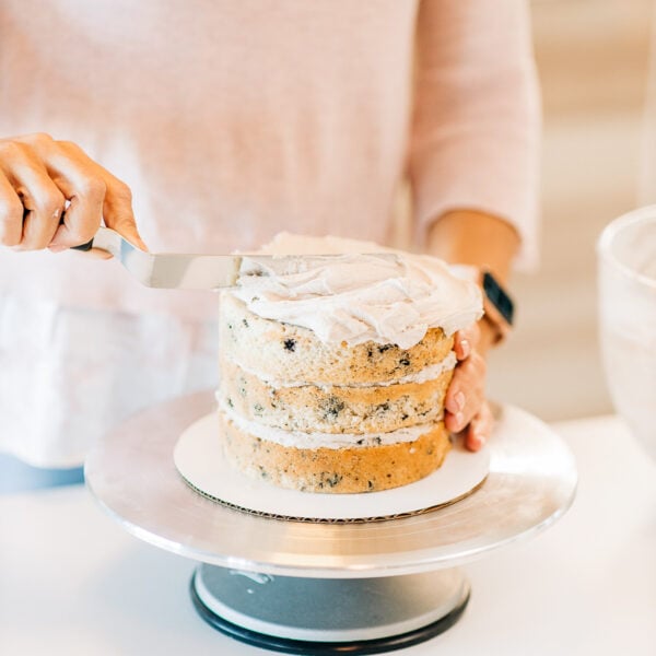 A person frosting a three-layer cake with a spatula on a cake stand, reflecting on the 7 things I wish I knew when starting egg-free baking.