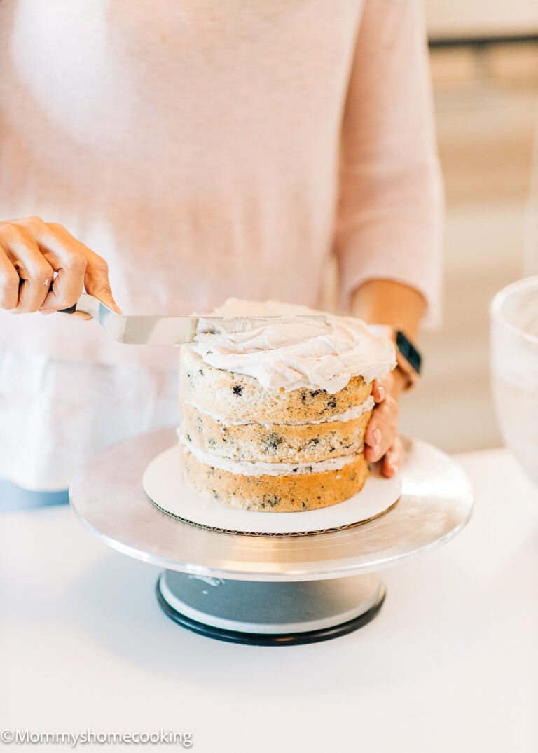 A person frosting a three-layer cake with a spatula on a cake stand, reflecting on the 7 things I wish I knew when starting egg-free baking.