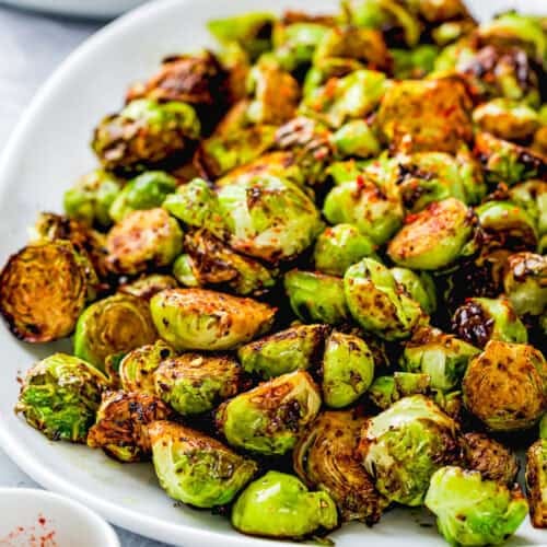 A white plate of roasted Brussels sprouts, lightly browned and perfectly crisped in the air fryer, garnished with red pepper flakes.