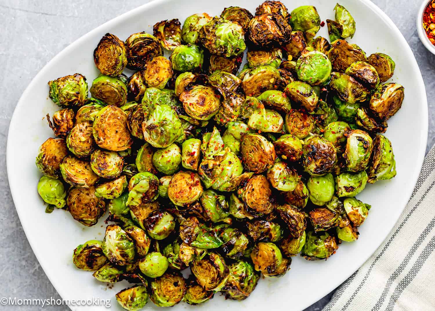An air fryer transforms Brussels sprouts into a savory dish, with charred and seasoned vegetables arranged on a white plate, set on a light gray surface next to a striped cloth.