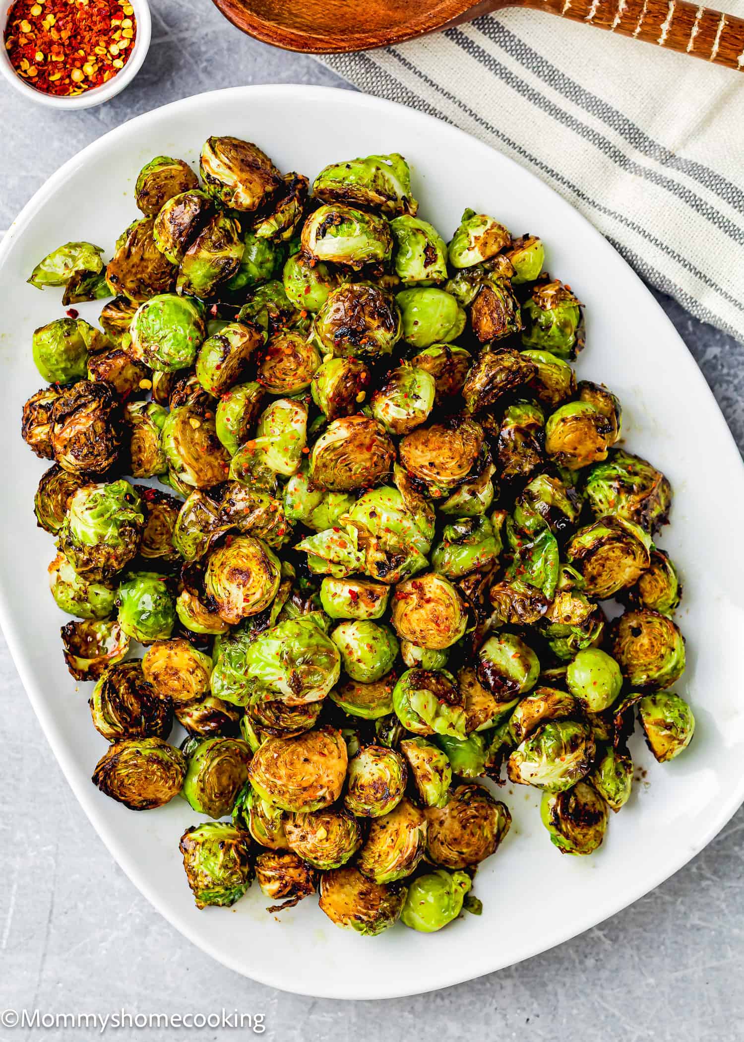 A white plate showcasing air fryer Brussels sprouts, perfectly roasted and slightly charred, with a bowl of red pepper flakes on the side.