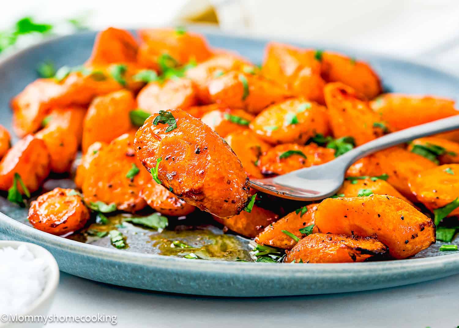 A fork holds a piece of roasted carrot above a plate filled with sliced, seasoned, and garnished Air Fryer Honey Glazed Carrots.