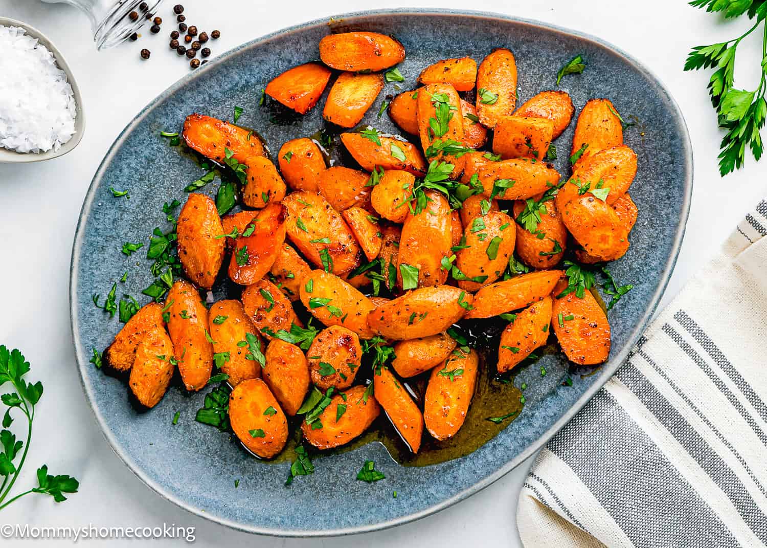 A gray plate filled with Air Fryer Honey Glazed Carrots garnished with chopped parsley, placed on a white surface with a striped napkin and fresh parsley nearby.
