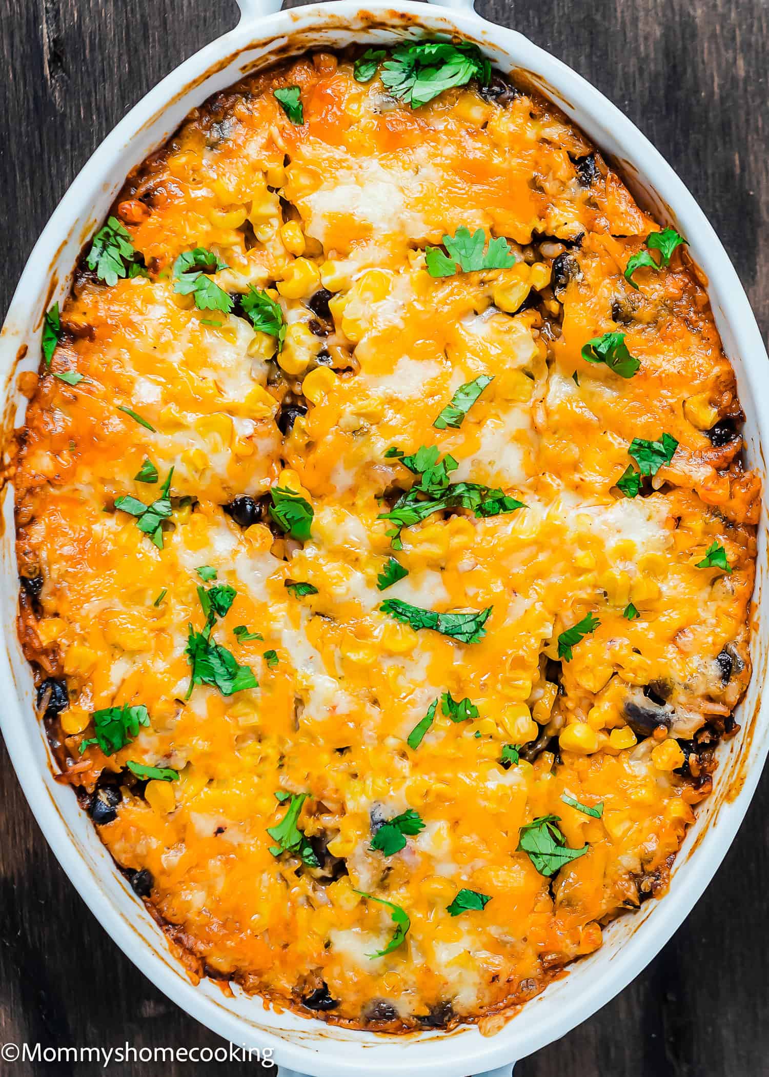 Cheesy Enchilada Rice and Beans Casserole baked with melted cheese, corn, black beans, and fresh cilantro, served in an oval white dish on a dark wooden surface.