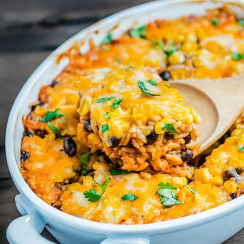 Cheesy Enchilada Rice and Beans Casserole topped with melted cheese, black beans, corn, and fresh cilantro is being served with a wooden spoon from a white dish.
