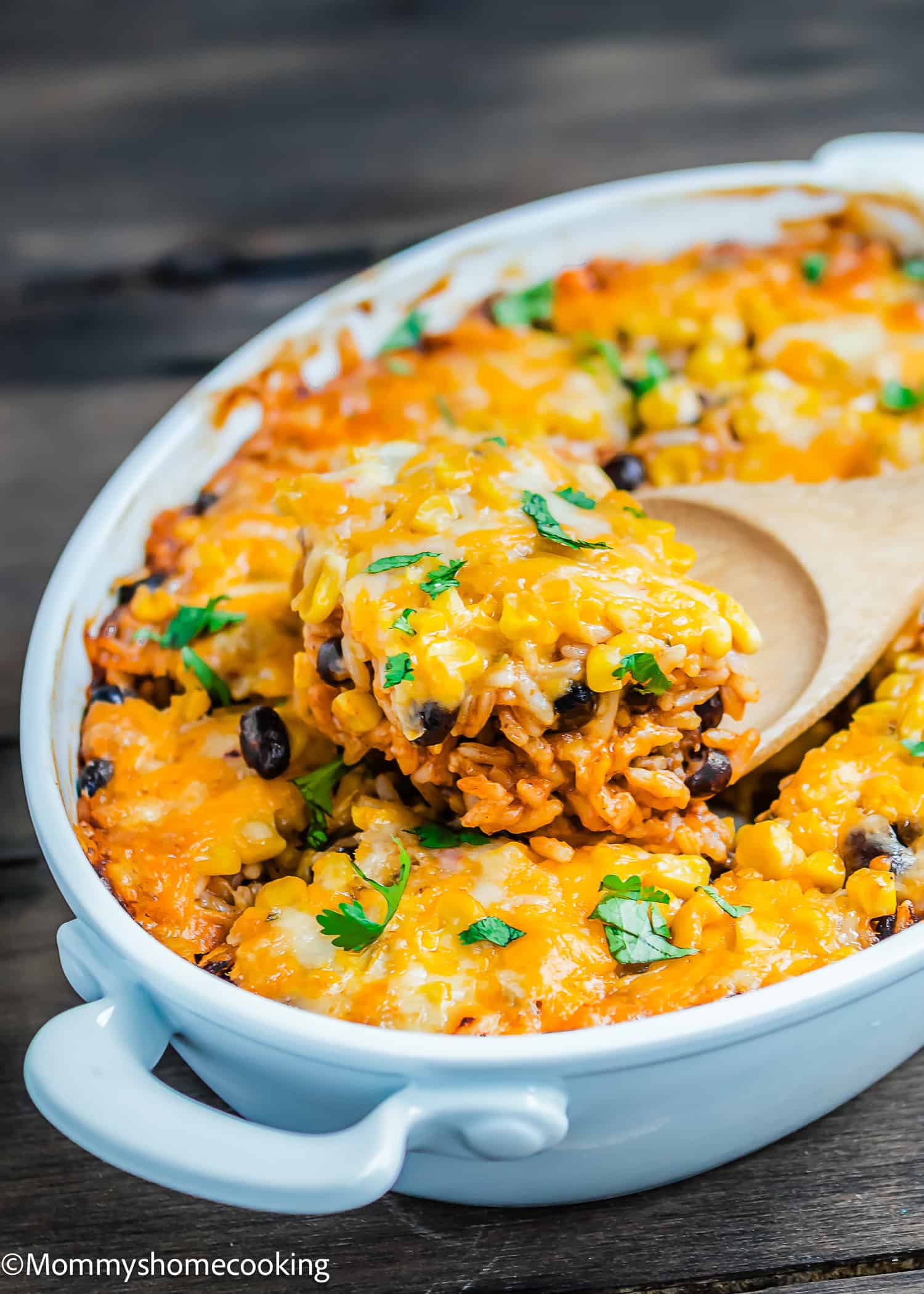 Cheesy Enchilada Rice and Beans Casserole topped with melted cheese, black beans, corn, and fresh cilantro is being served with a wooden spoon from a white dish.