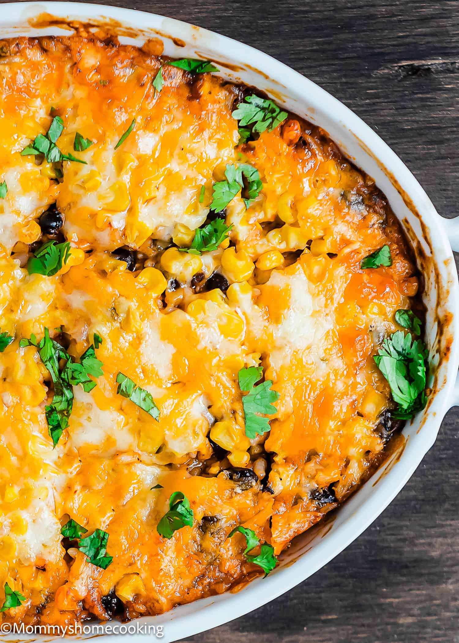 A Cheesy Enchilada Rice and Beans Casserole topped with melted cheese, corn, black beans, and garnished with chopped cilantro in a white dish on a wooden surface.