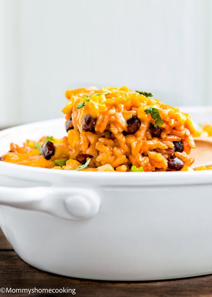A serving of Cheesy Enchilada Rice and Beans Casserole with black beans and corn in a white ceramic dish, garnished with chopped herbs.