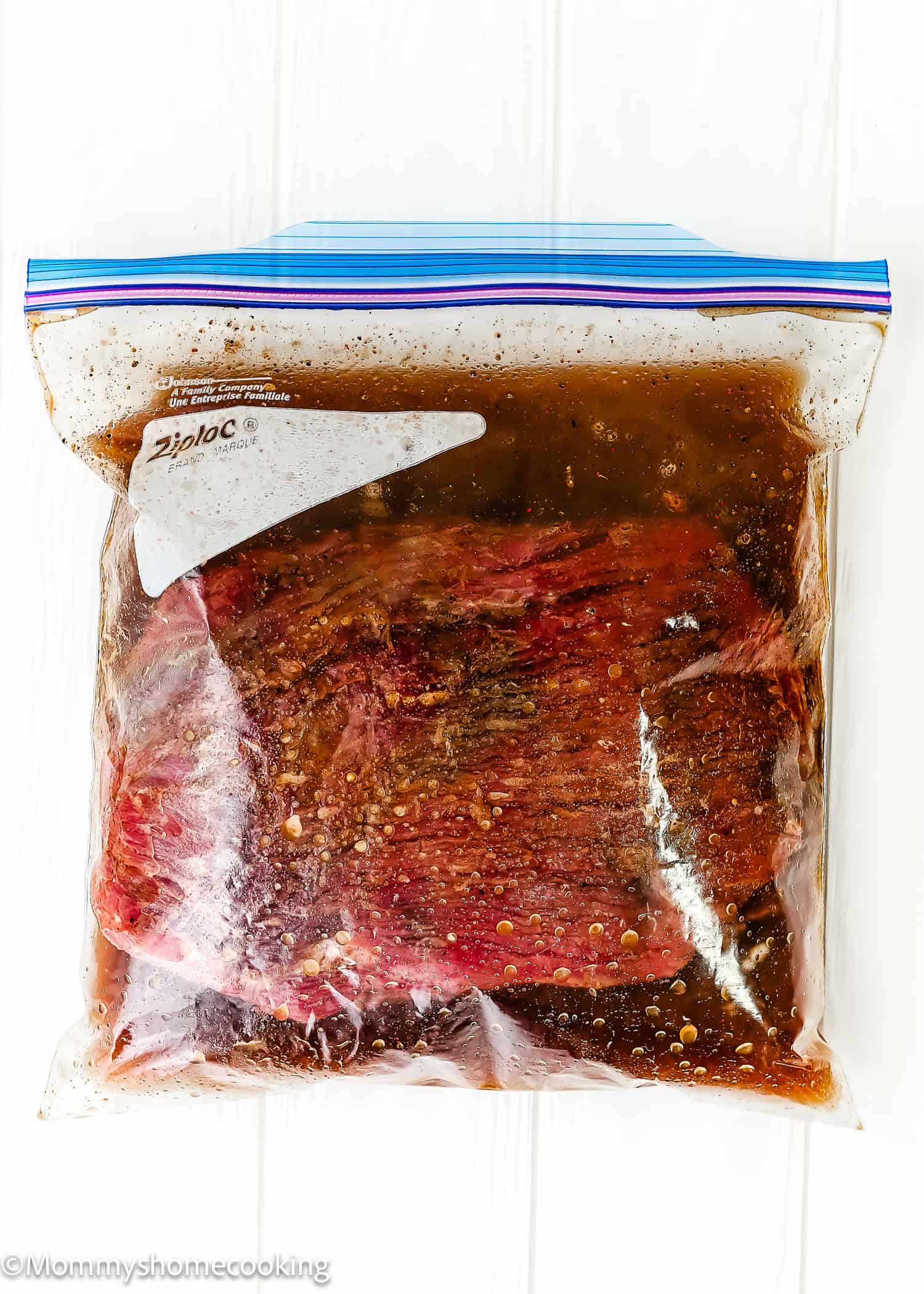 Easy Carne Asada with the best marinade ever: a large piece of raw meat marinating in a sealed plastic zip-top bag filled with dark liquid on a white surface.