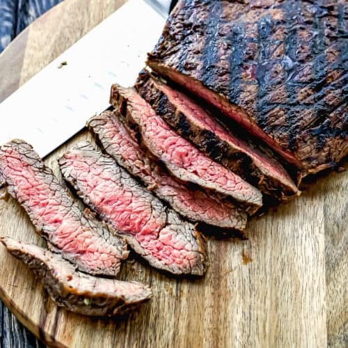 Easy Carne Asada with the best marinade ever—sliced grilled steak with a seared exterior and pink center is arranged on a wooden cutting board beside a large knife.