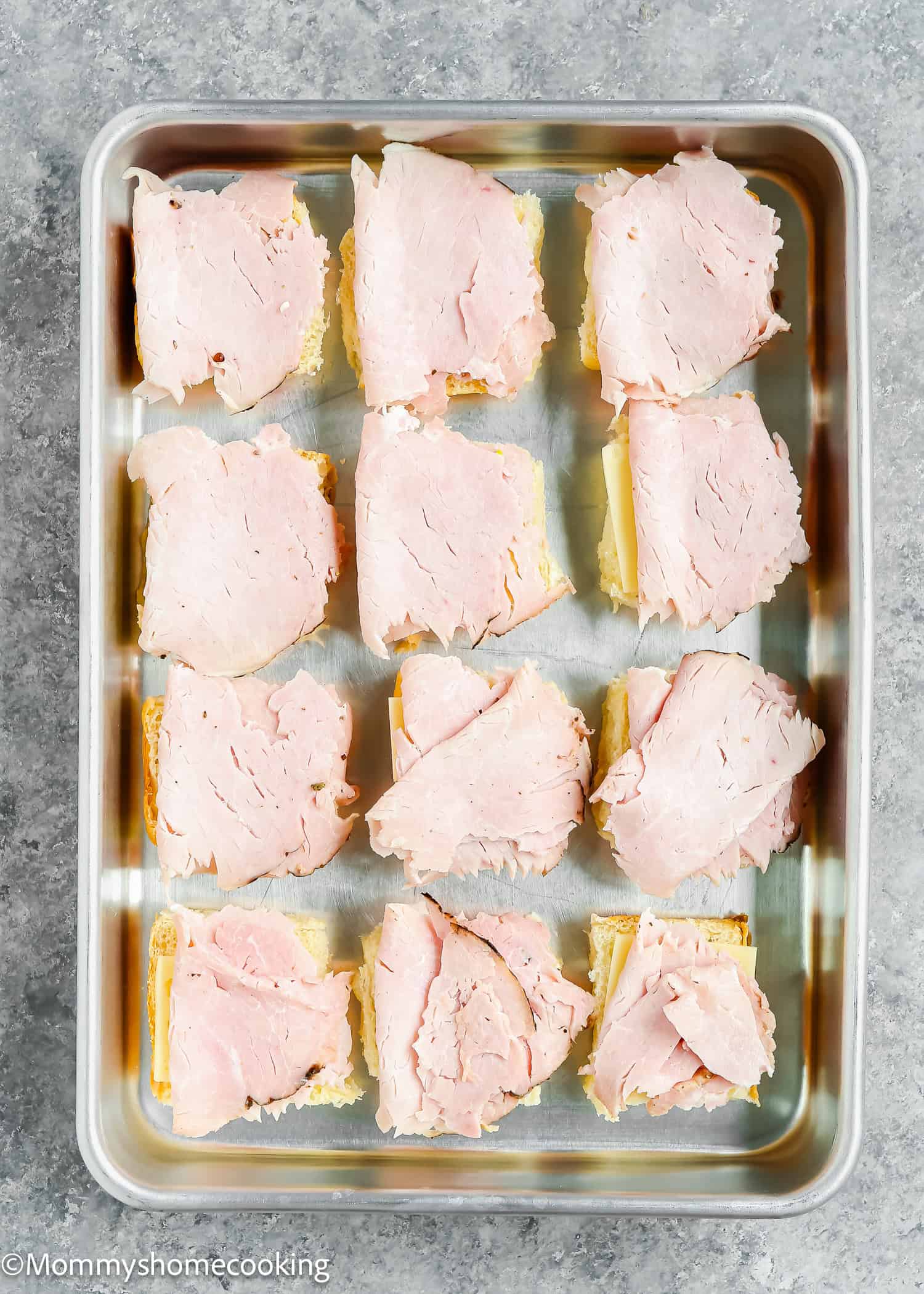Twelve pieces of bread topped with sliced ham are arranged in a metal baking tray on a gray surface, ready to become easy Cuban sliders. Ready in 30 minutes!.