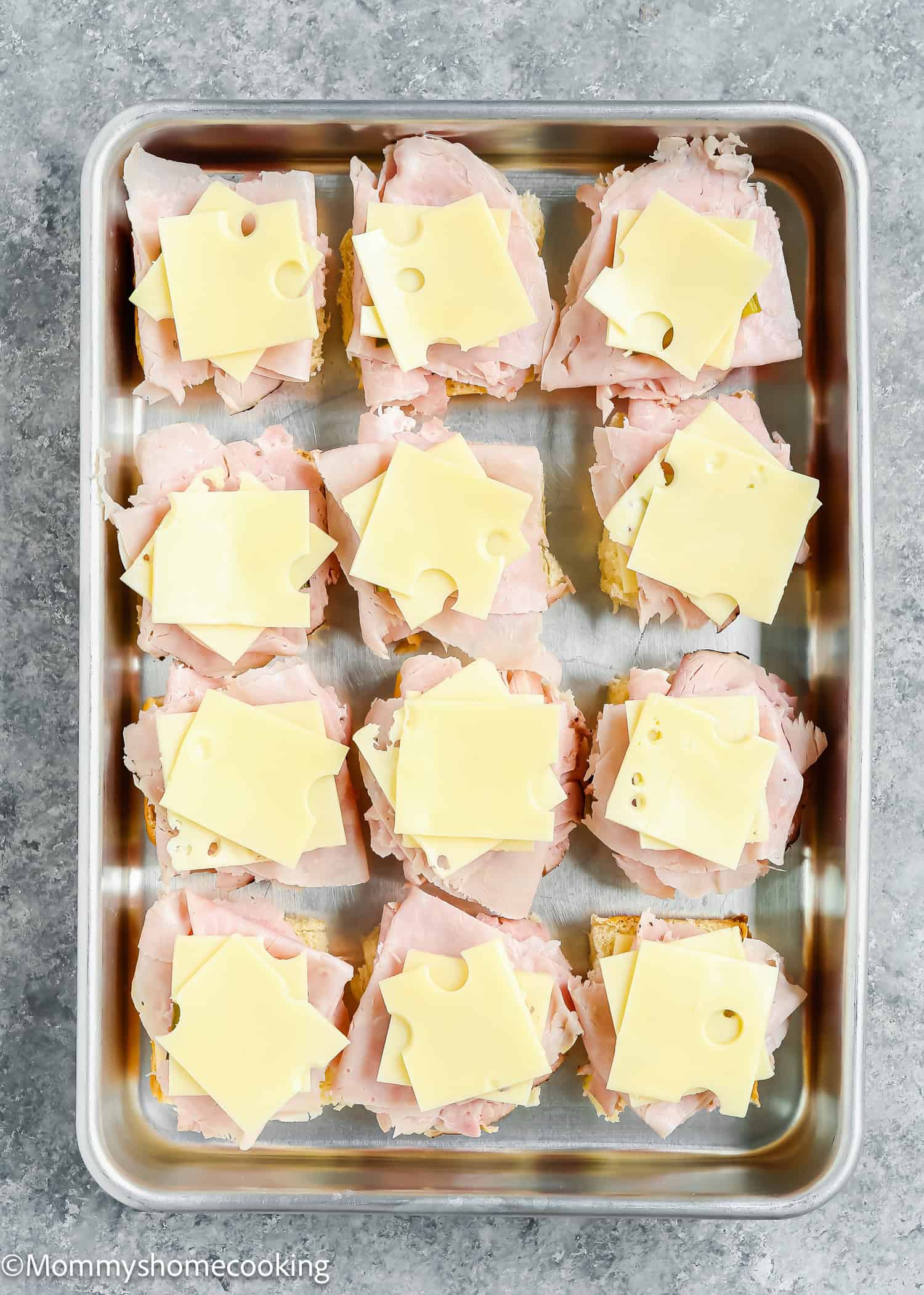 Easy Cuban Sliders ready in 30 minutes are arranged on a rectangular tray, each topped with sliced ham and Swiss cheese, placed on a textured gray surface.