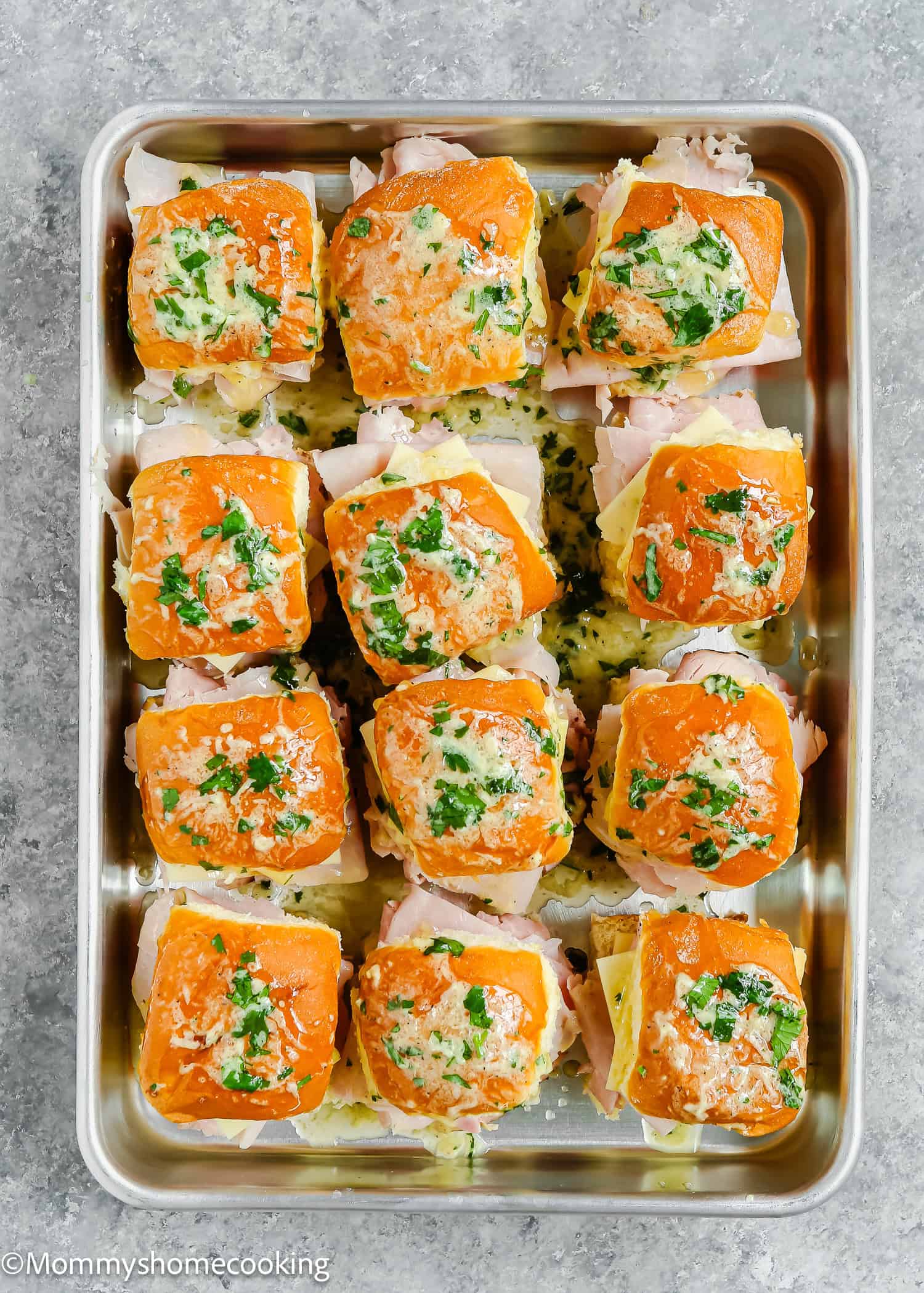 Easy Cuban Sliders, ready in 30 minutes, featuring twelve baked sliders with ham and melted cheese. They're topped with herbs and butter and perfectly arranged in a metal baking tray.