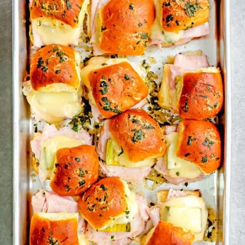 Easy Cuban sliders, bursting with flavors and topped with a buttery herb glaze, are ready in 30 minutes. Arranged neatly on a metal baking tray, these baked ham and cheese delights offer the perfect blend of taste and convenience for any gathering.