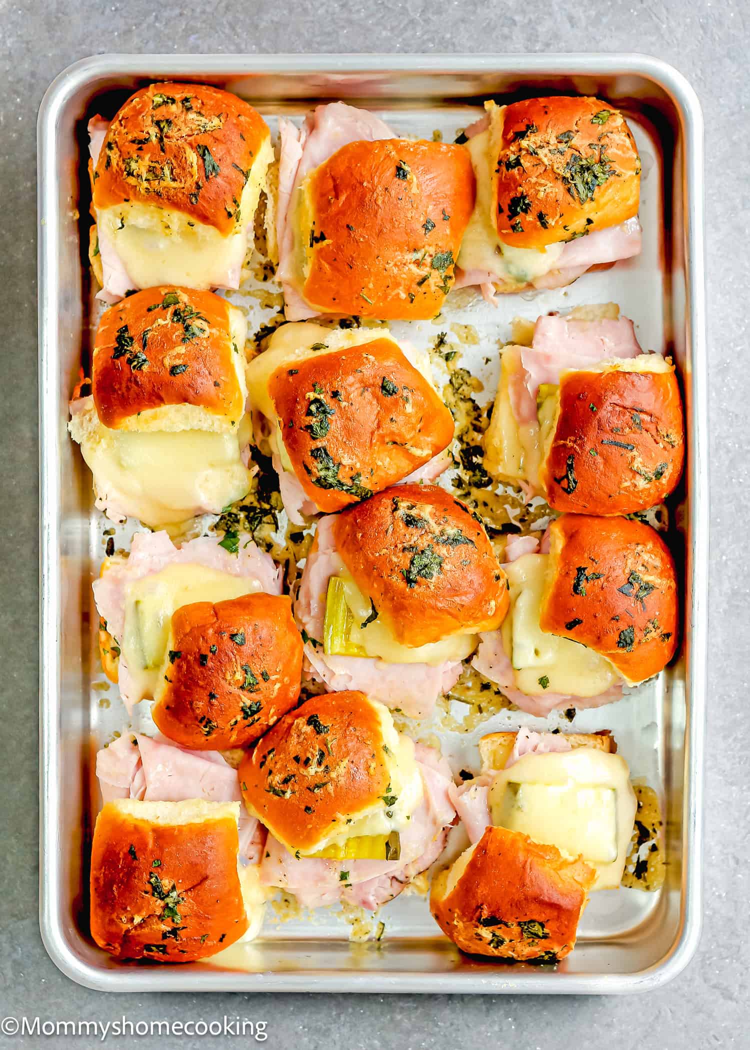 Easy Cuban sliders, bursting with flavors and topped with a buttery herb glaze, are ready in 30 minutes. Arranged neatly on a metal baking tray, these baked ham and cheese delights offer the perfect blend of taste and convenience for any gathering.