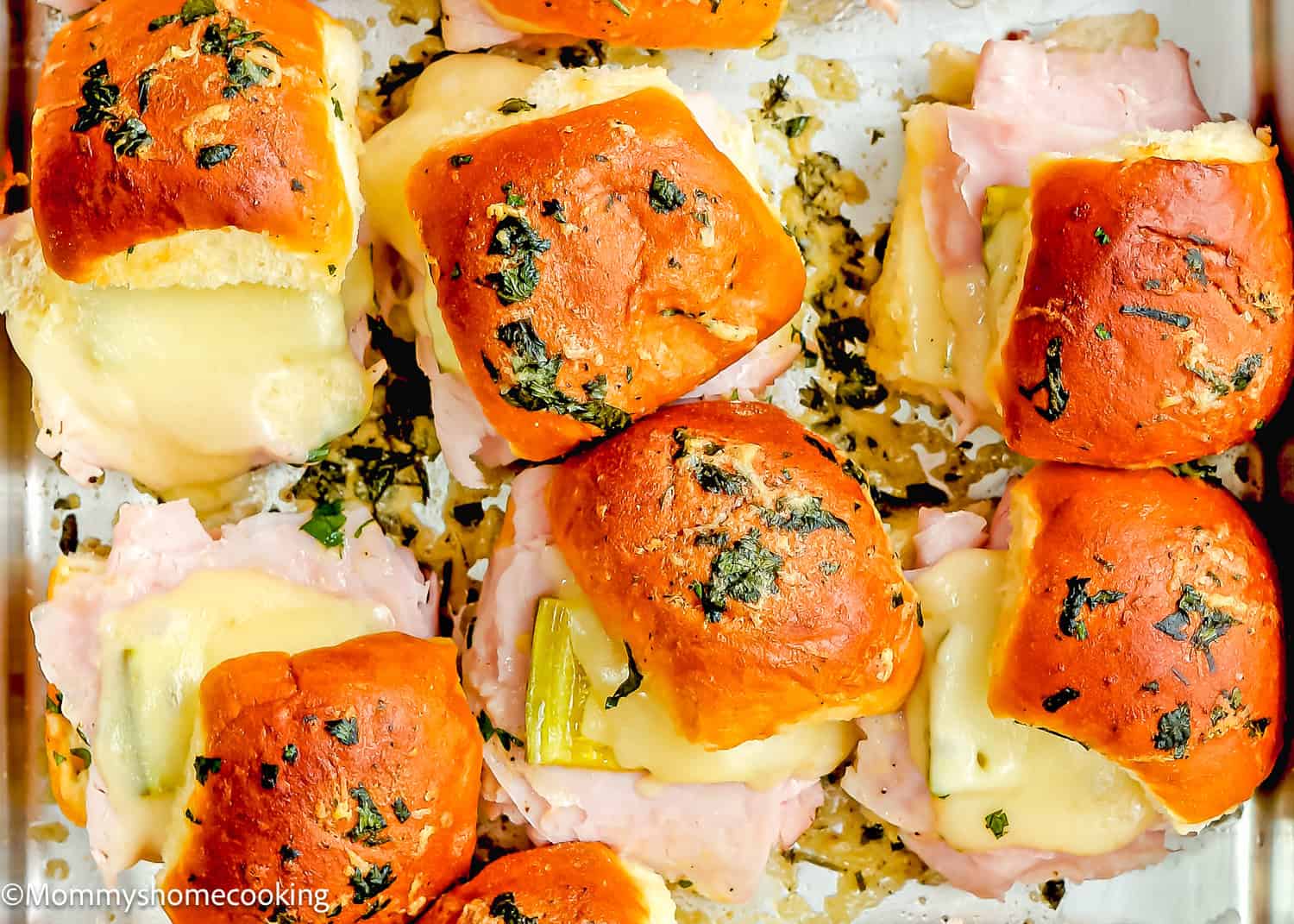 Easy Cuban Sliders, ready in just 30 minutes, feature a close-up of baked ham and cheese topped with melted cheese and sprinkled with chopped herbs on a baking tray.