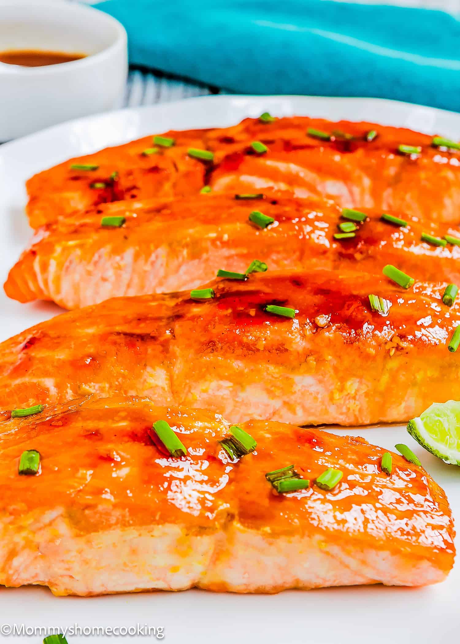 Three Easy Glazed Salmon fillets topped with chopped chives are arranged on a white plate, accompanied by a lime wedge and a small bowl of sauce in the background.