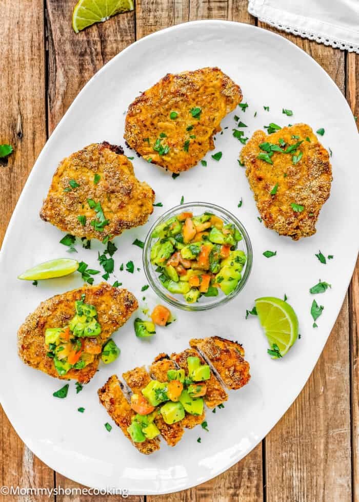 Five breaded chicken pieces on a white plate, garnished with chopped herbs, served with a bowl of avocado salsa and lime wedges—perfect for fans of Oven Fried Pork Chops seeking something new.