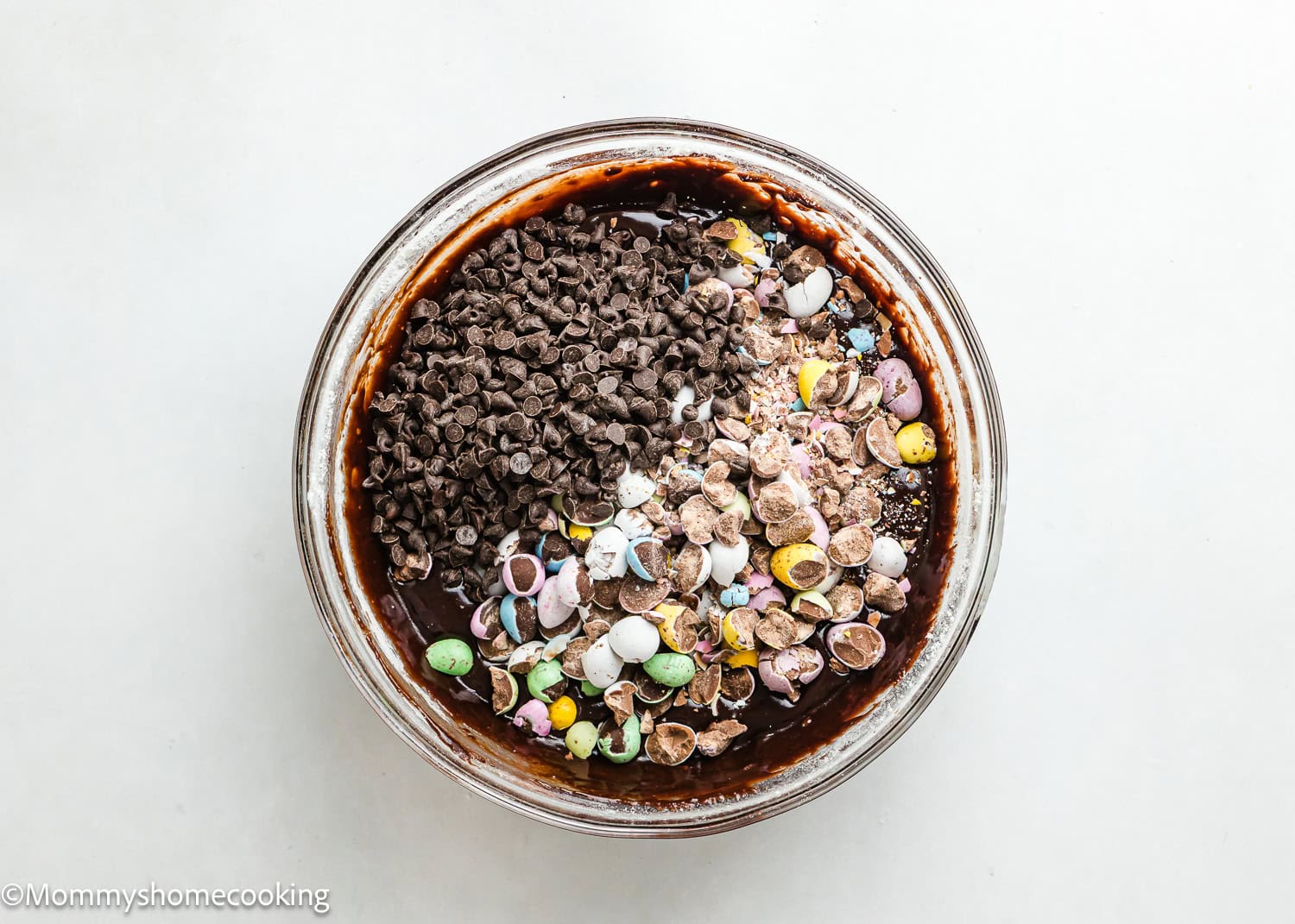 A glass bowl filled with eggless brownie batter, topped with crushed colorful candies, chopped chocolate, Cadbury Mini Eggs, and chocolate chips.