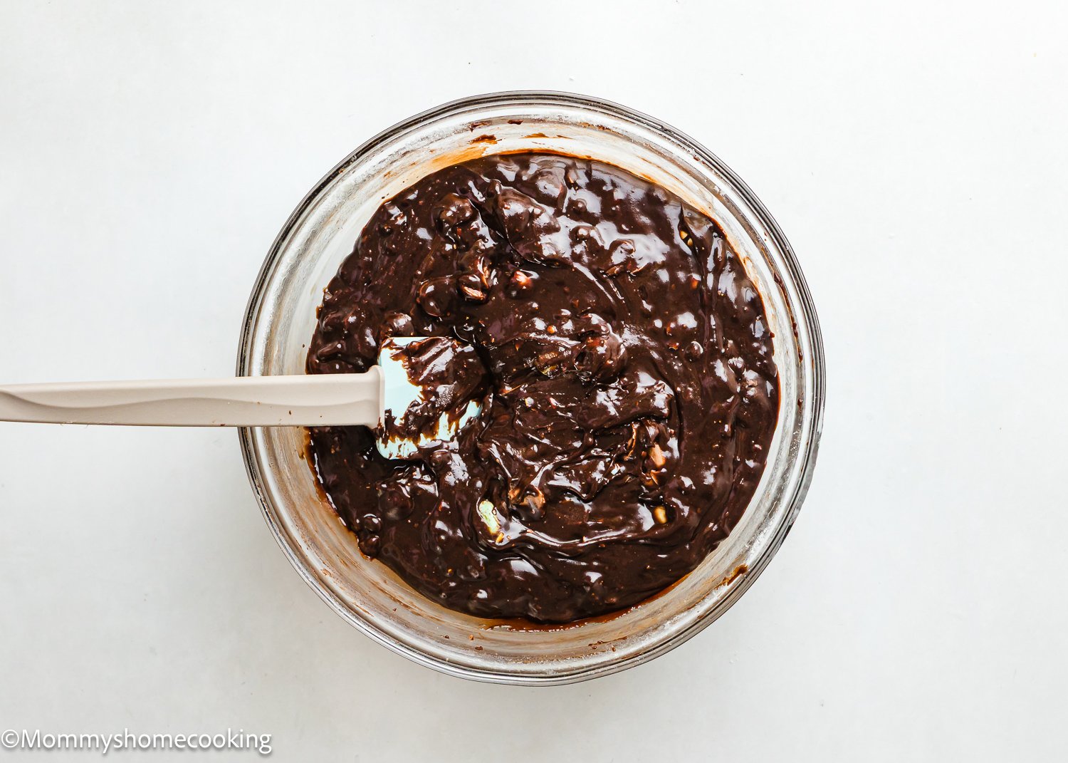 A glass bowl brimming with chocolate batter and a white spatula on a light background, perfect for whipping up Eggless Cadbury Mini Egg Brownies.