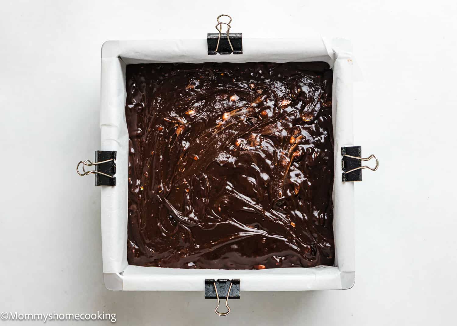 A square baking pan lined with parchment paper, filled with eggless Cadbury Mini Egg brownie batter, secured with binder clips on the edges.