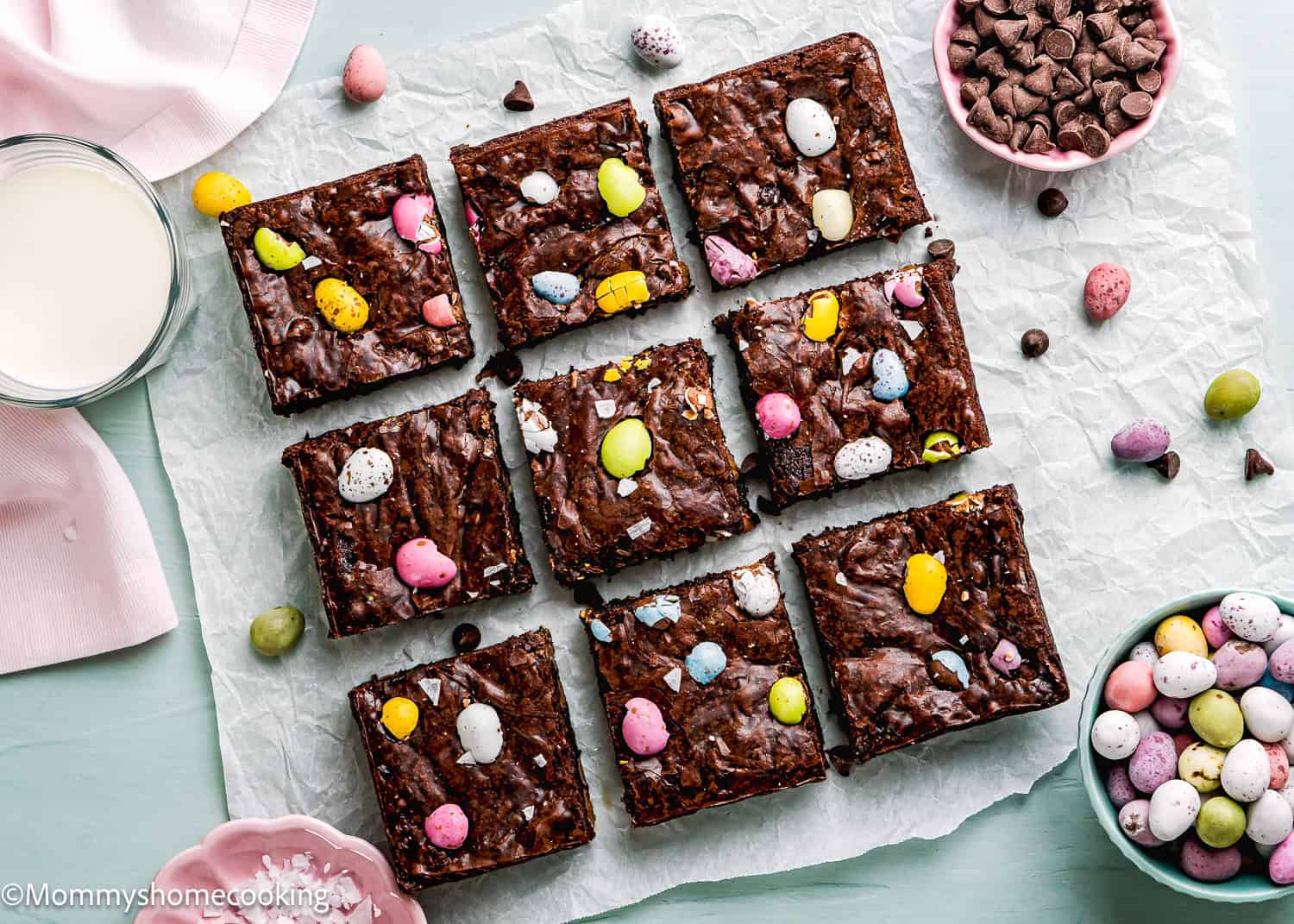 Arrayed on parchment paper are nine eggless Cadbury Mini Egg brownies, each topped with vibrant candy eggs. Beside them sit a glass of milk and bowls of chocolate chips and candy eggs on a light blue surface.