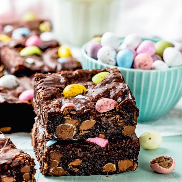Eggless brownies with colorful candy pieces are stacked beside a bowl of Cadbury Mini Eggs and a glass of milk on a light blue surface.