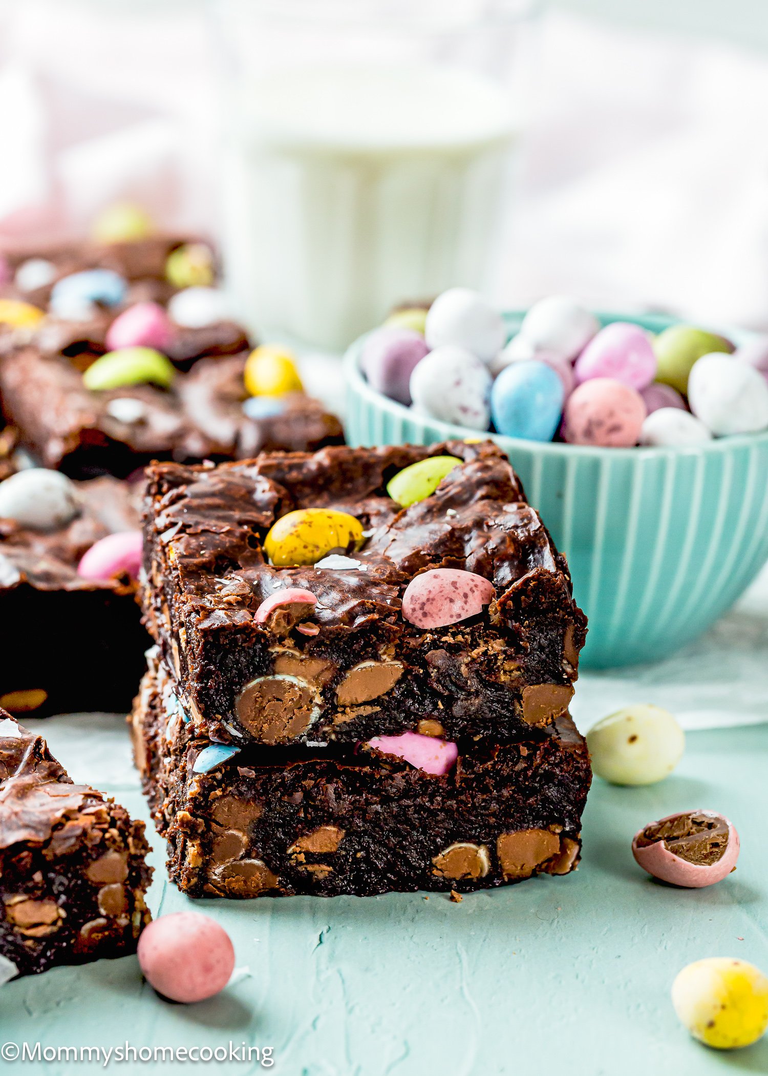 Eggless brownies with colorful candy pieces are stacked beside a bowl of Cadbury Mini Eggs and a glass of milk on a light blue surface.