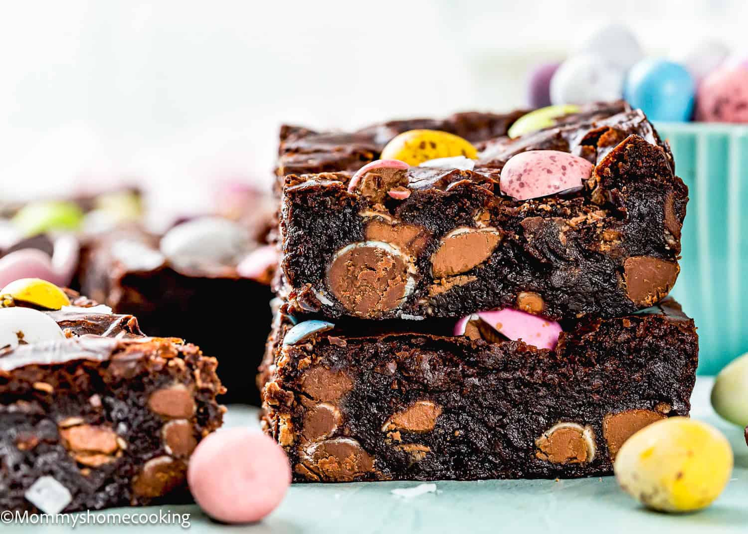 Three stacked eggless brownies with colorful candy pieces, Cadbury Mini Eggs, and chocolate chips, surrounded by an assortment of candies.