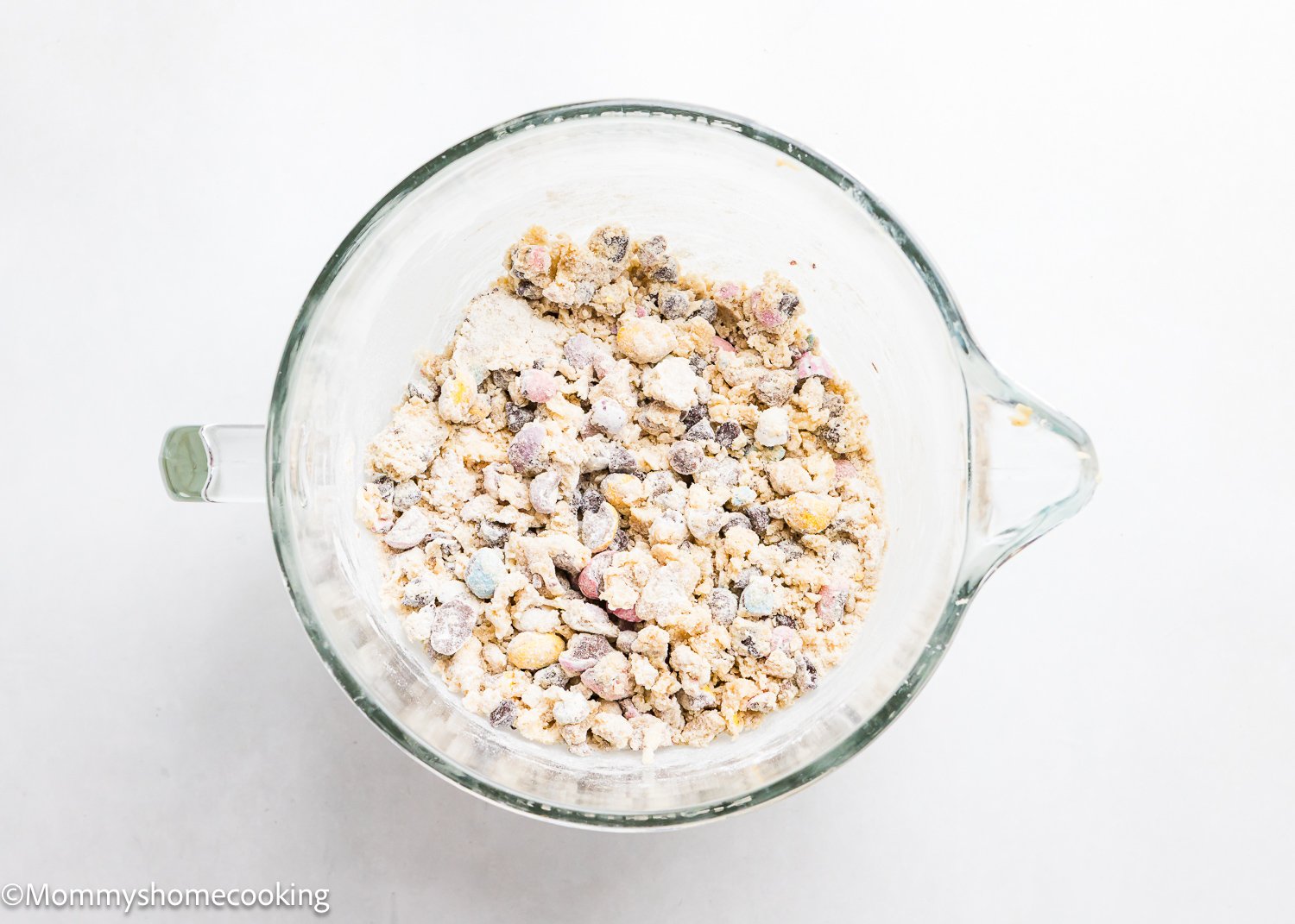 A glass bowl holds an assortment of dry ingredients and small pieces, perfect for crafting eggless Cadbury Mini Egg cookies, all resting on a pristine white surface.