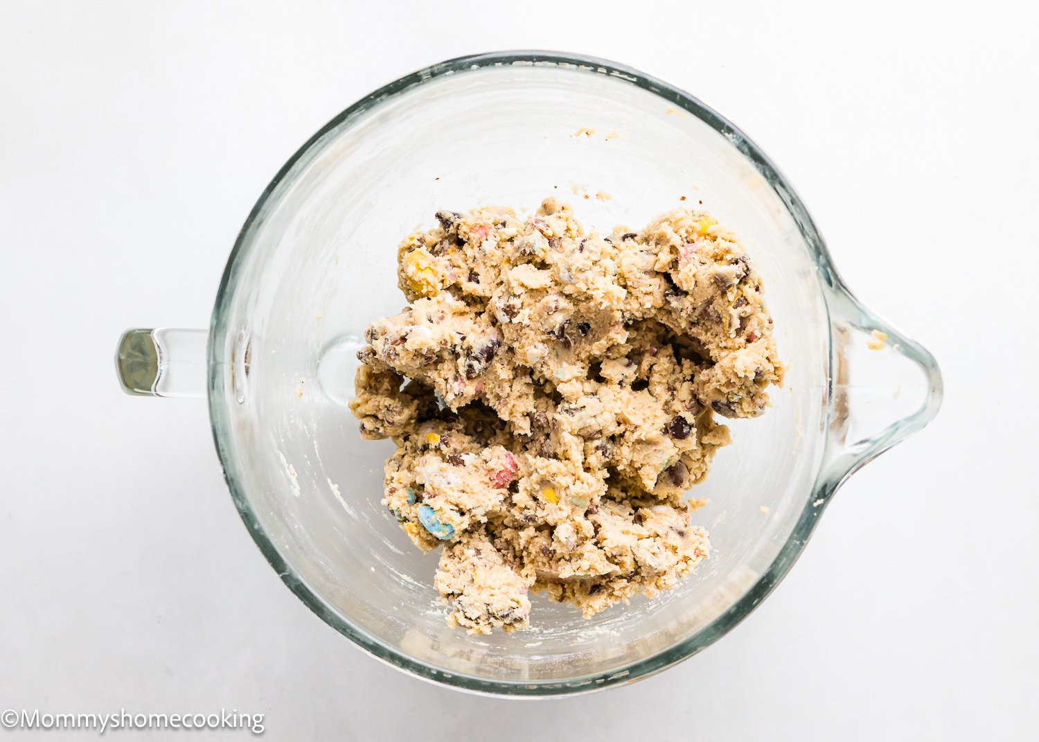A glass bowl filled with eggless cookie dough mixed with chocolate chips and Cadbury Mini Eggs on a white background.