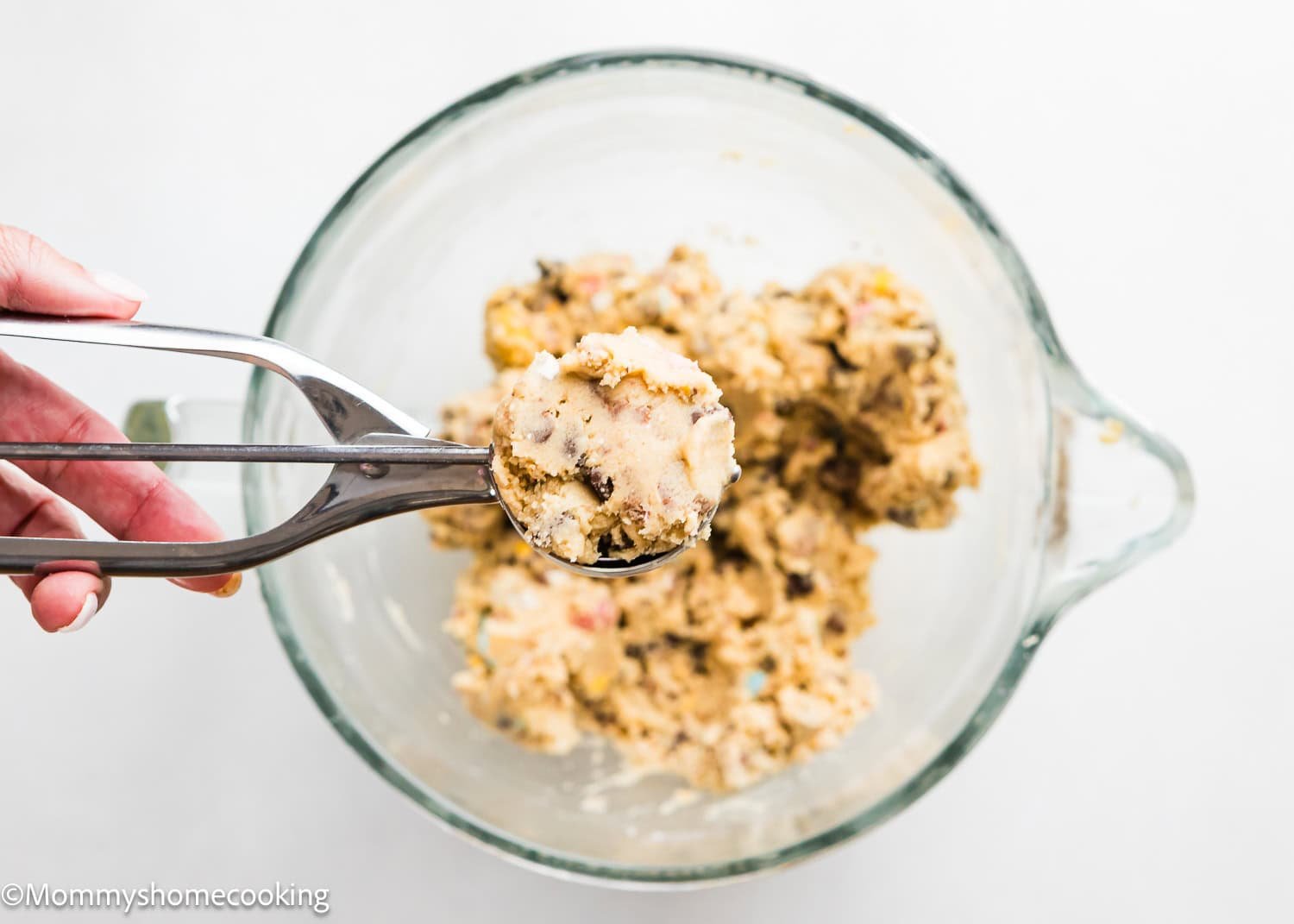 A hand holds a metal scoop of eggless cookie dough above a glass bowl filled with more dough, featuring delightful Cadbury Mini Eggs, set against a plain white background.
