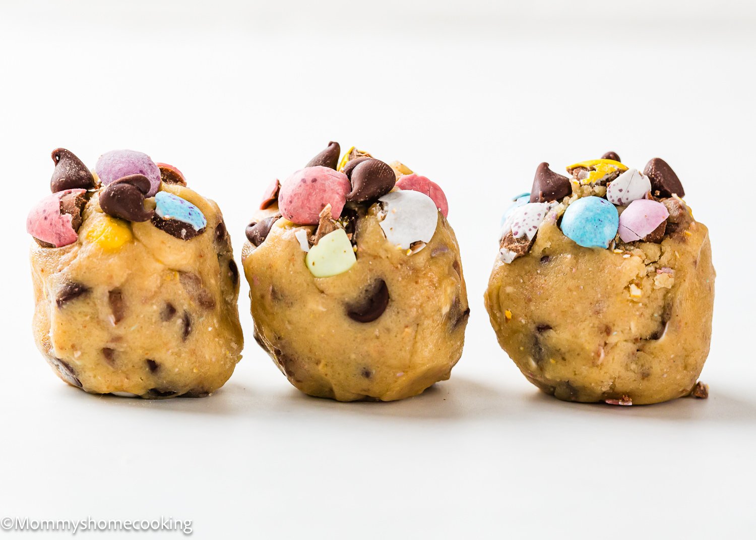Three balls of eggless cookie dough topped with colorful Cadbury Mini Eggs and chocolate chips on a white background.