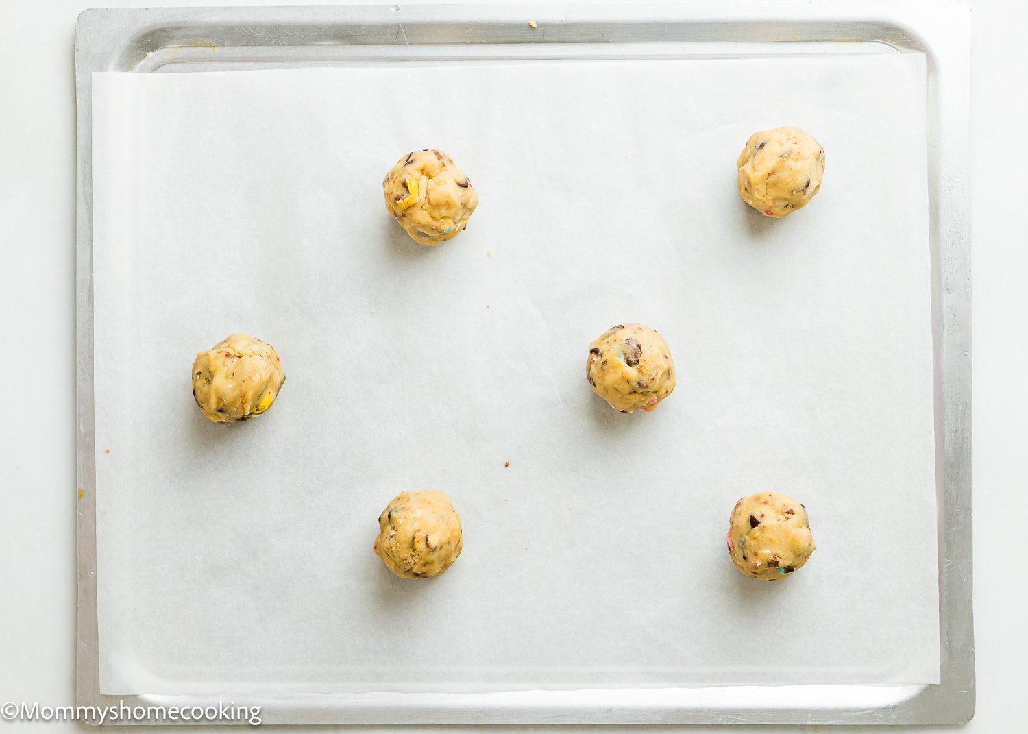 Six eggless Cadbury Mini Egg cookie dough balls rest on a parchment-lined baking tray.