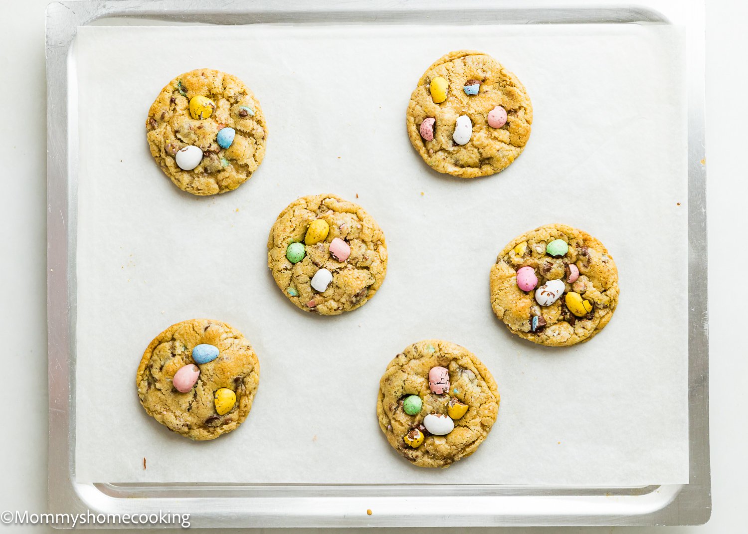 A baking tray with six Eggless Cadbury Mini Egg Cookies is placed on white parchment paper, each cookie topped with vibrant candy-coated chocolate eggs.