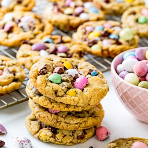 A stack of eggless cookies with colorful candy-coated chocolates and chocolate chips sits on a cooling rack, next to a bowl of Cadbury Mini Eggs.