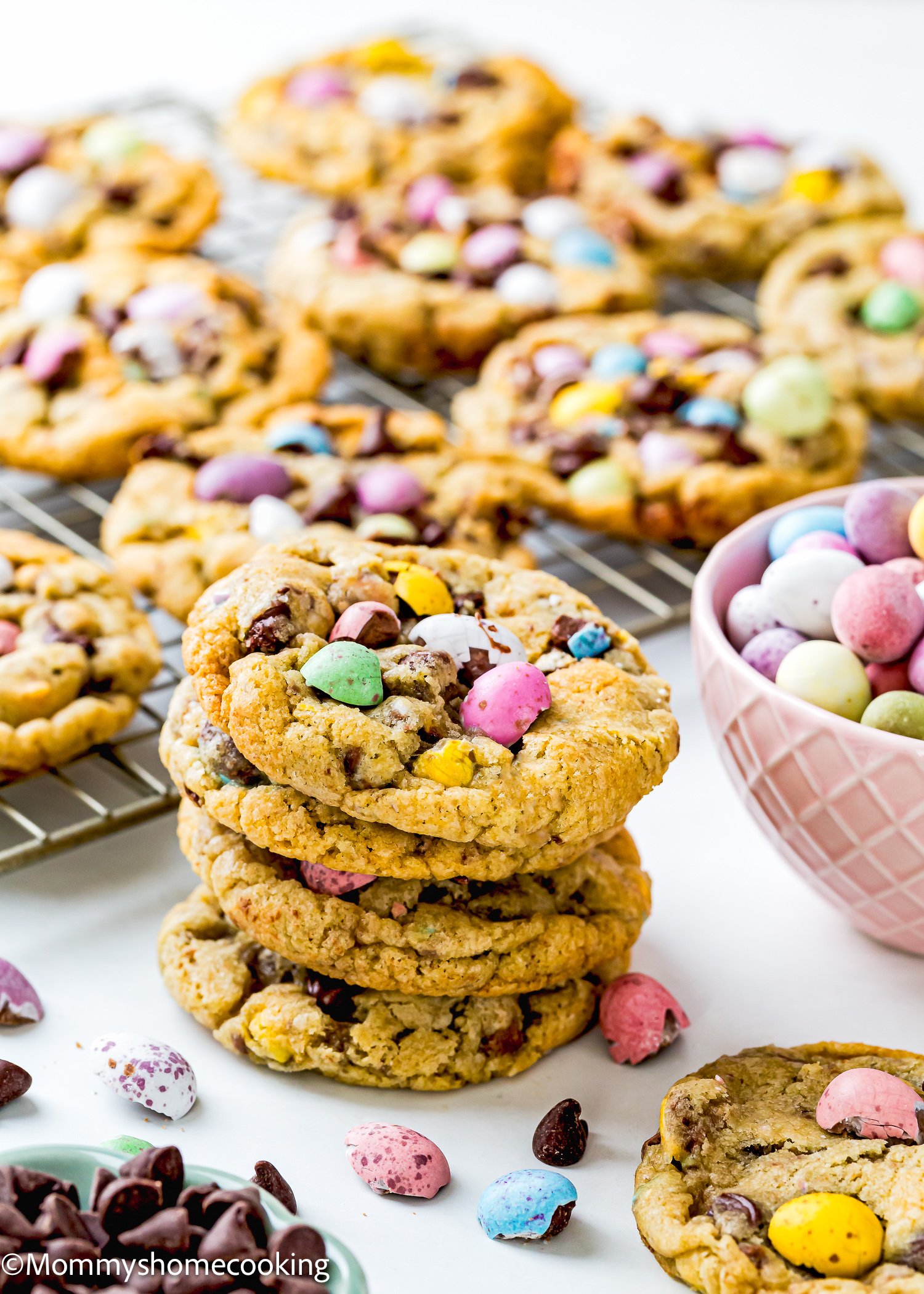 A stack of eggless cookies with colorful candy-coated chocolates and chocolate chips sits on a cooling rack, next to a bowl of Cadbury Mini Eggs.