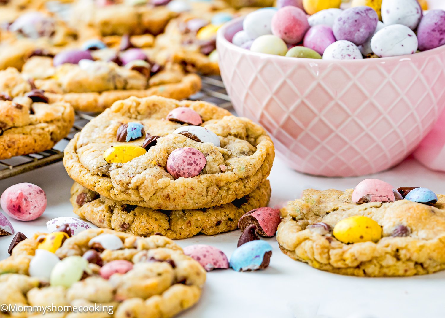 Eggless Cadbury Mini Egg Cookies adorned with colorful candy-coated chocolates sit next to a bowl brimming with similar treats.