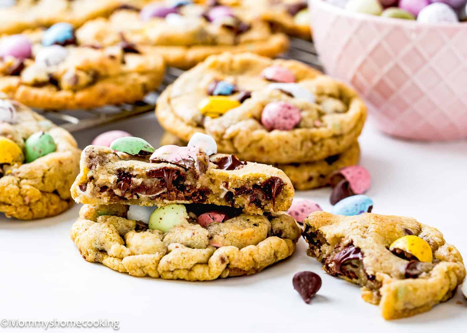 Eggless cookies with colorful candy-coated chocolates and chocolate chips rest on a white surface. One cookie is broken in half, revealing melting chocolate inside. A pink bowl filled with candies adds a pop of color in the background.