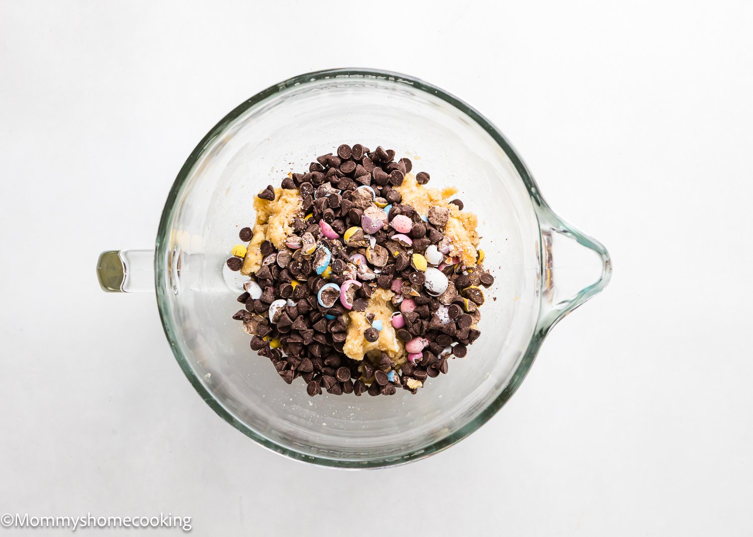 A glass bowl filled with cookie dough, chocolate chips, and colorful candies sits on a white background, perfect for making eggless Cadbury Mini Egg Cookies.