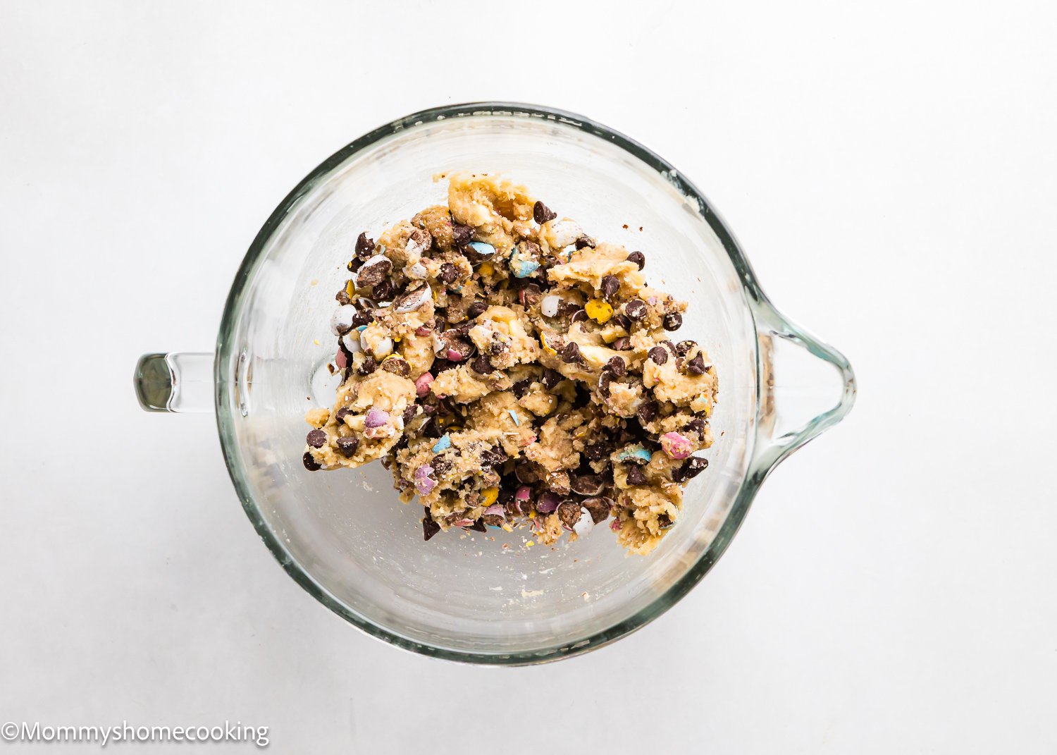 An eggless glass bowl brimming with cookie dough, mixed with chocolate chips and colorful Cadbury Mini Egg pieces, sits on a pristine white surface.