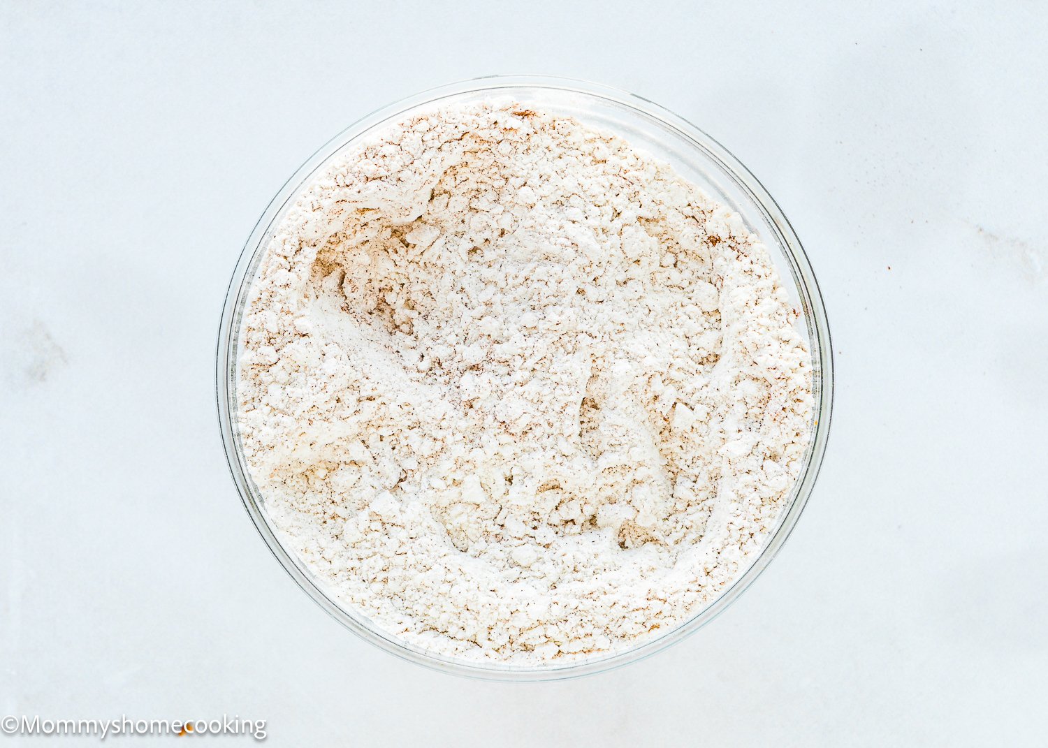 A glass bowl filled with white and light brown dry flour mixture sits on a light surface, perfect for making eggless carrot banana muffins.