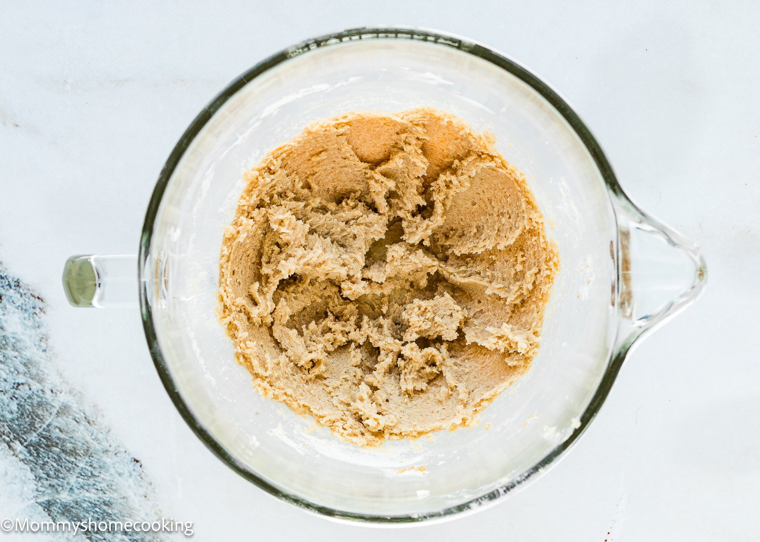 A clear mixing bowl holds creamy brown batter for eggless carrot banana muffins, placed on a light surface.
