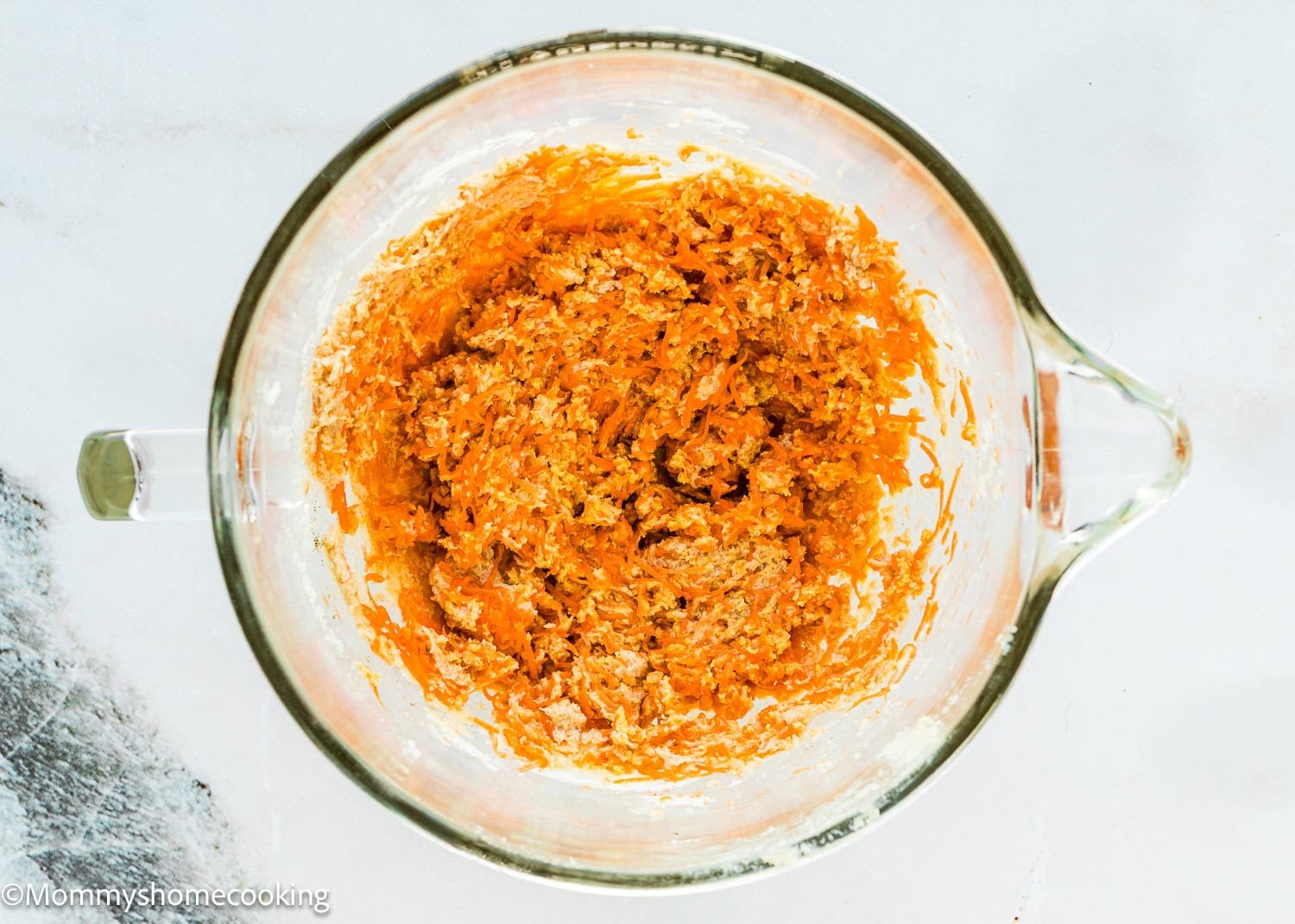 A glass bowl sits on a white countertop, filled with a mixed orange and brown shredded substance, ideal for making Eggless Carrot Banana Muffins.