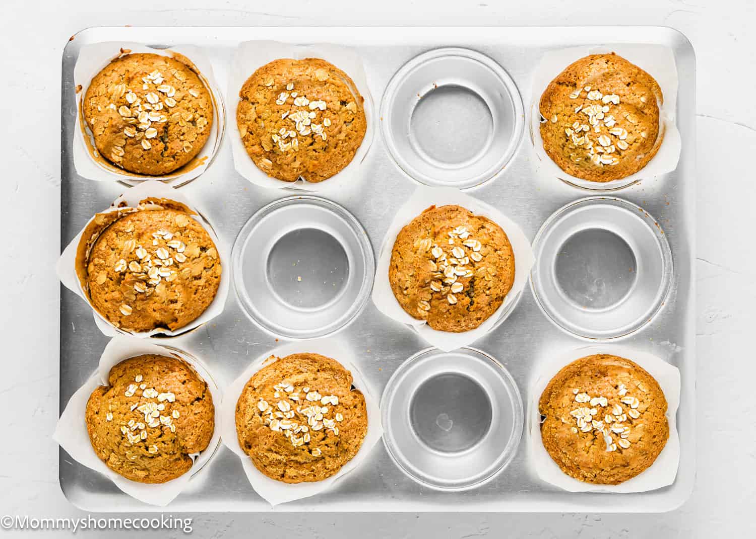 A muffin tray with six freshly baked eggless carrot banana muffins and four empty spaces on a white surface.