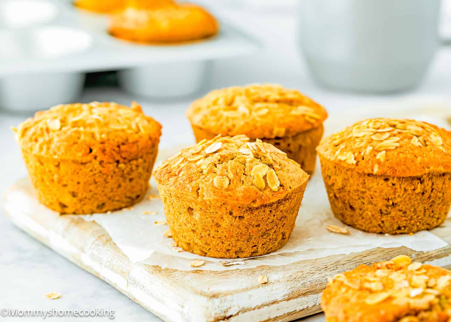 Eggless Carrot Banana Muffins, golden brown and topped with oats, are artfully arranged on a white wooden board, with a muffin tray in the background.